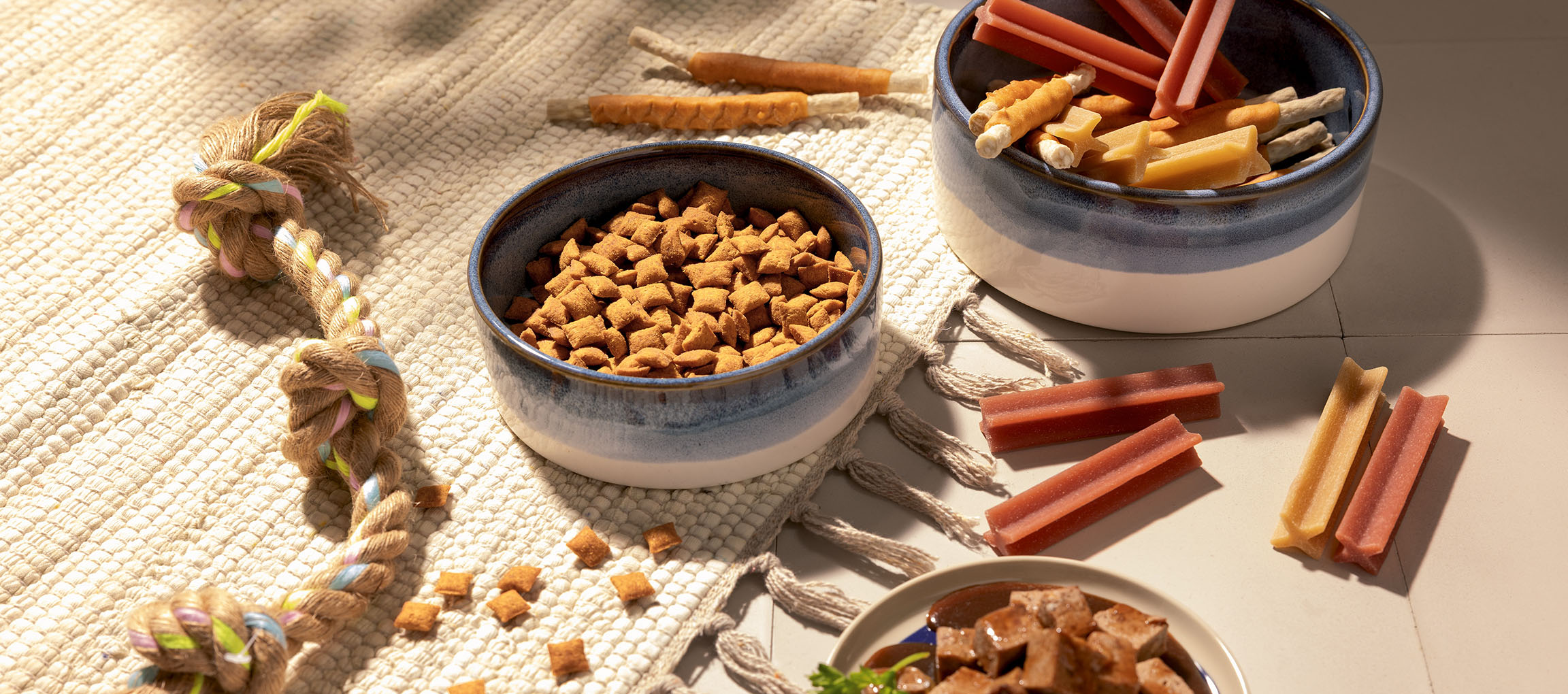 Alimentos e petiscos variados para animais de estimação, incluindo ração seca, paus de mastigar e um brinquedo de corda.
