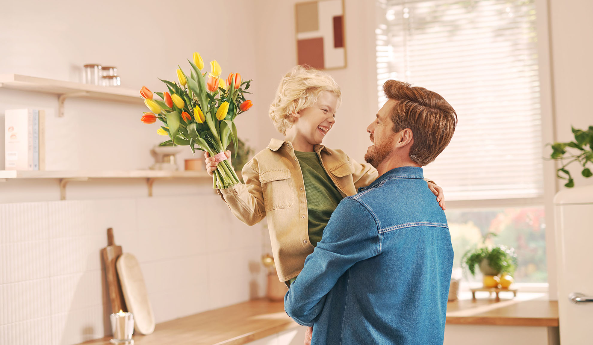 Pai a segurar o filho que oferece um ramo de tulipas numa cozinha luminosa.