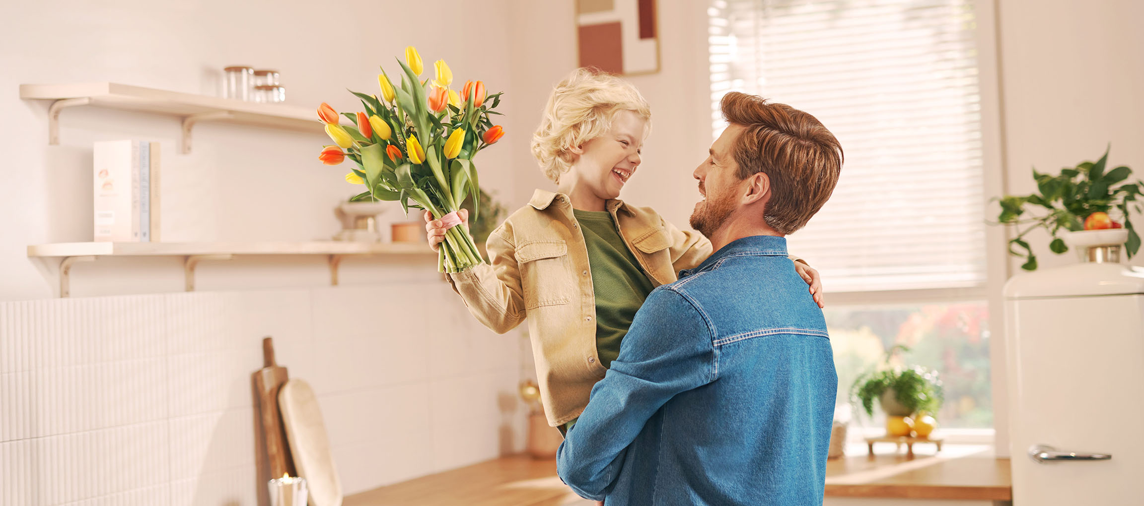 Um menino segurando um buquê de tulipas, rindo com um homem na cozinha.