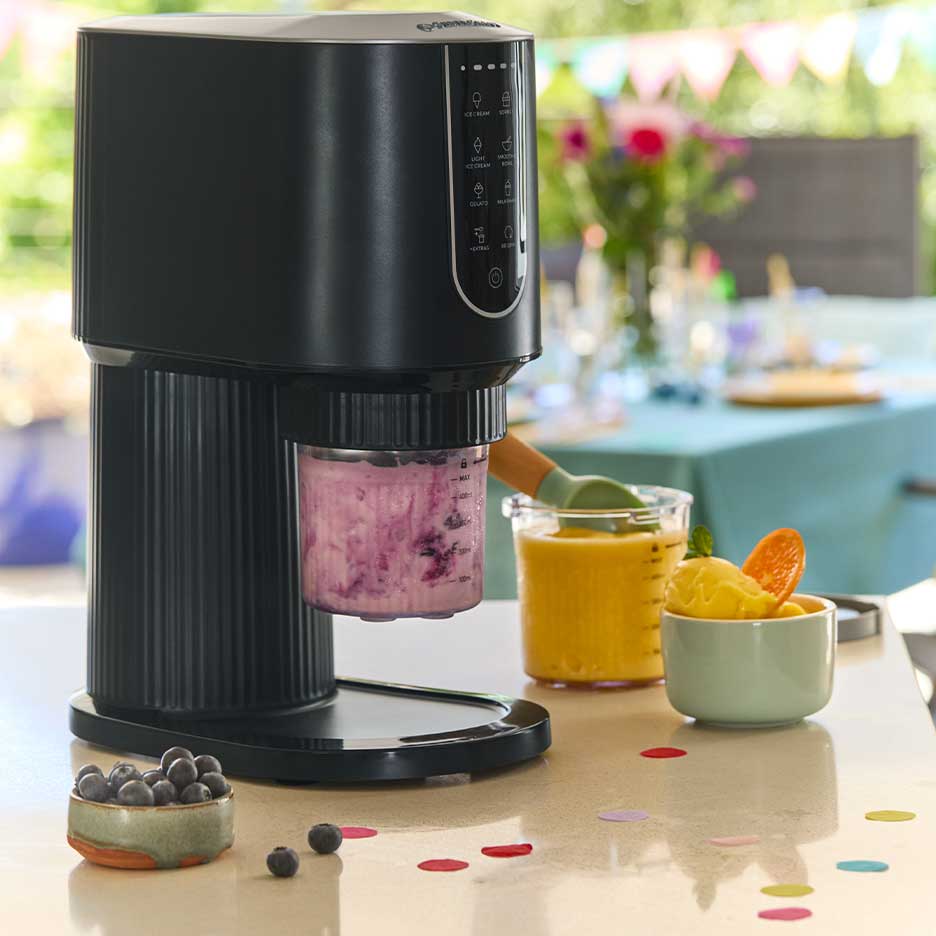 Máquina de gelados preta com gelado roxo, rodeada por gelado de manga e mirtilos.
