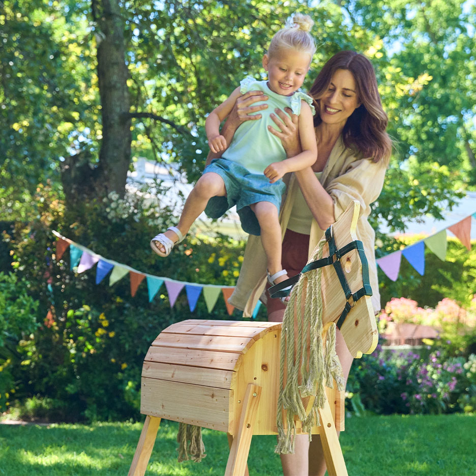 Mulher e menina a brincar com um cavalo de baloiço de madeira num jardim com bandeirinhas.