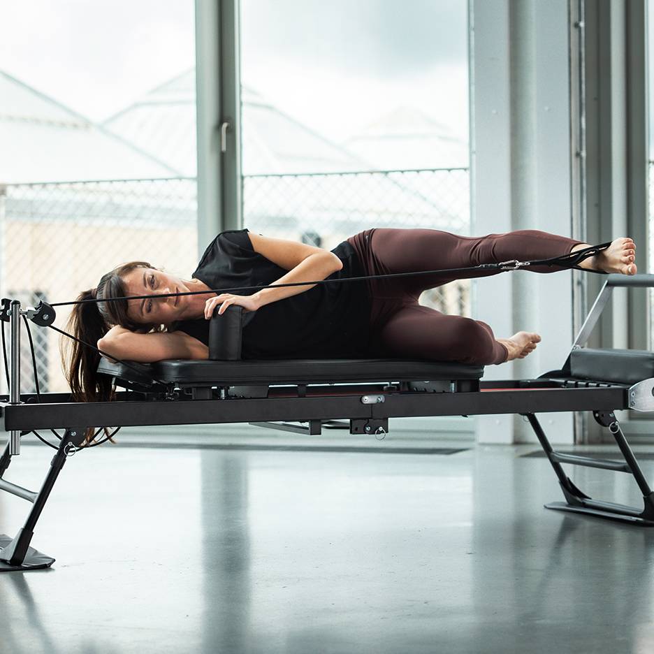 Mulher a fazer Pilates numa máquina reformer, deitada de lado com as correias nas pernas.