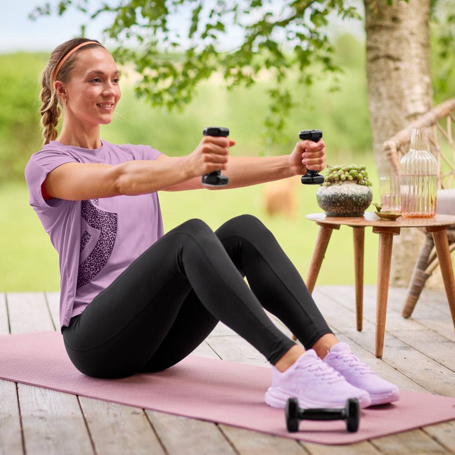 Mulher de t-shirt roxa e leggings pretas a exercitar-se com halteres num tapete de ioga.