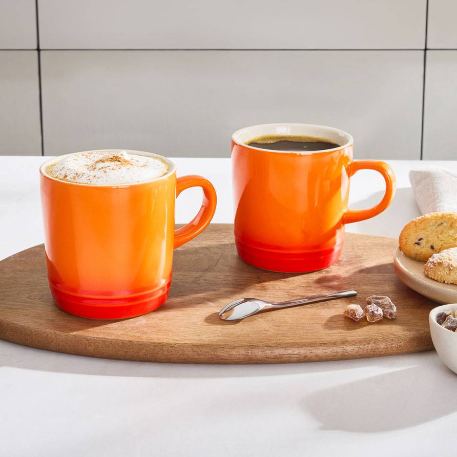 Duas canecas de café laranja numa bandeja de madeira, uma com café espumoso, a outra com café preto.