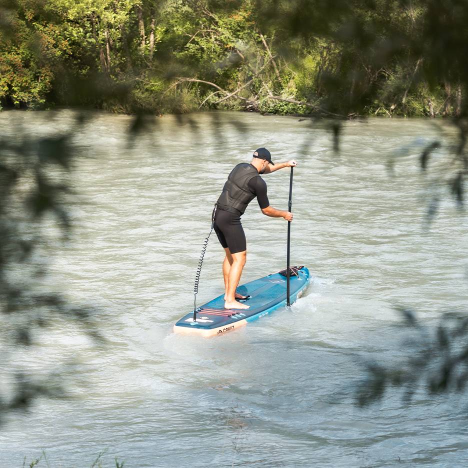 Homem em fato de mergulho e colete de flutuação a remar numa prancha de paddleboard azul num rio.