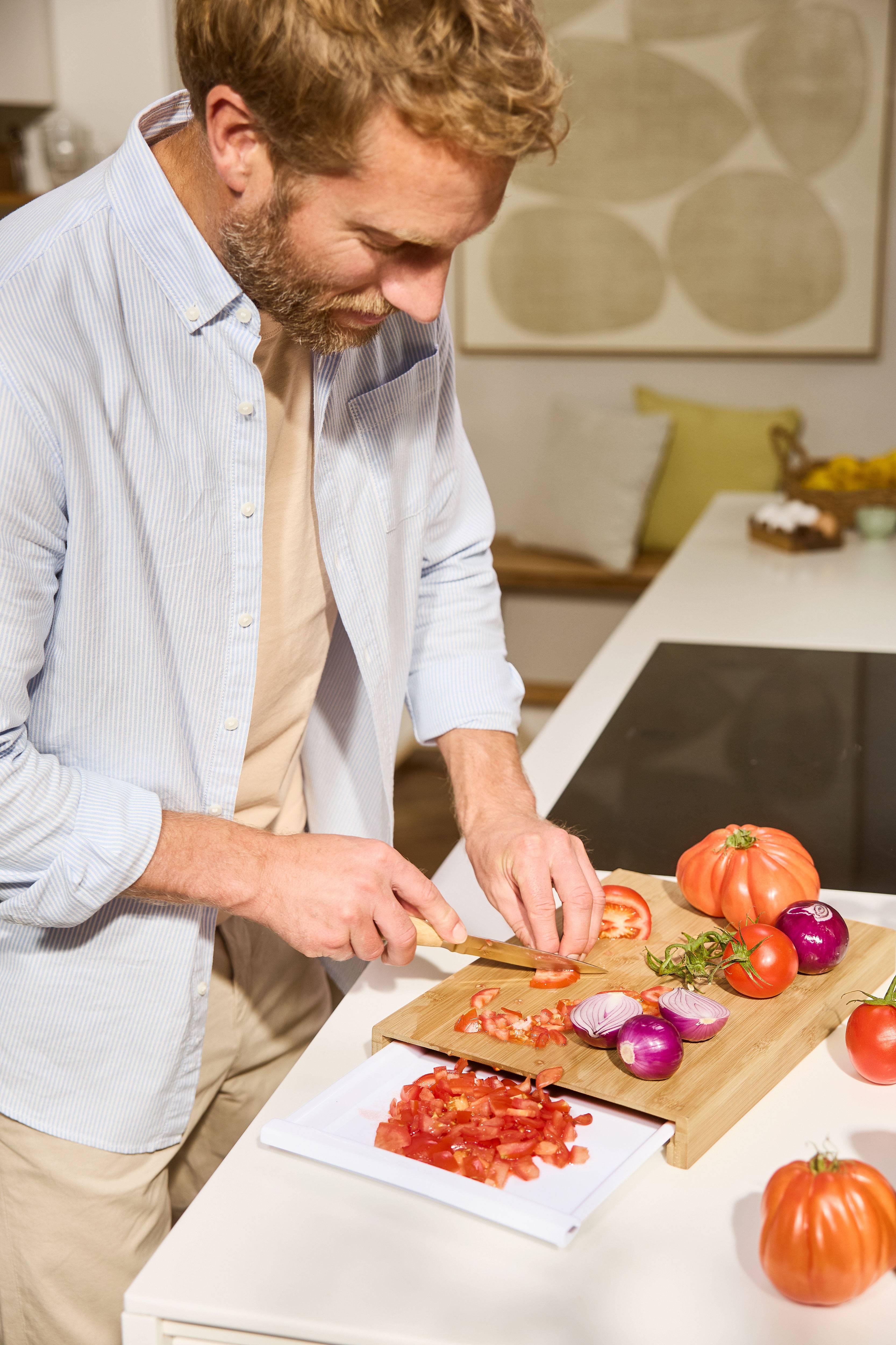 Homem a cortar tomates e cebolas numa tábua de madeira na cozinha.