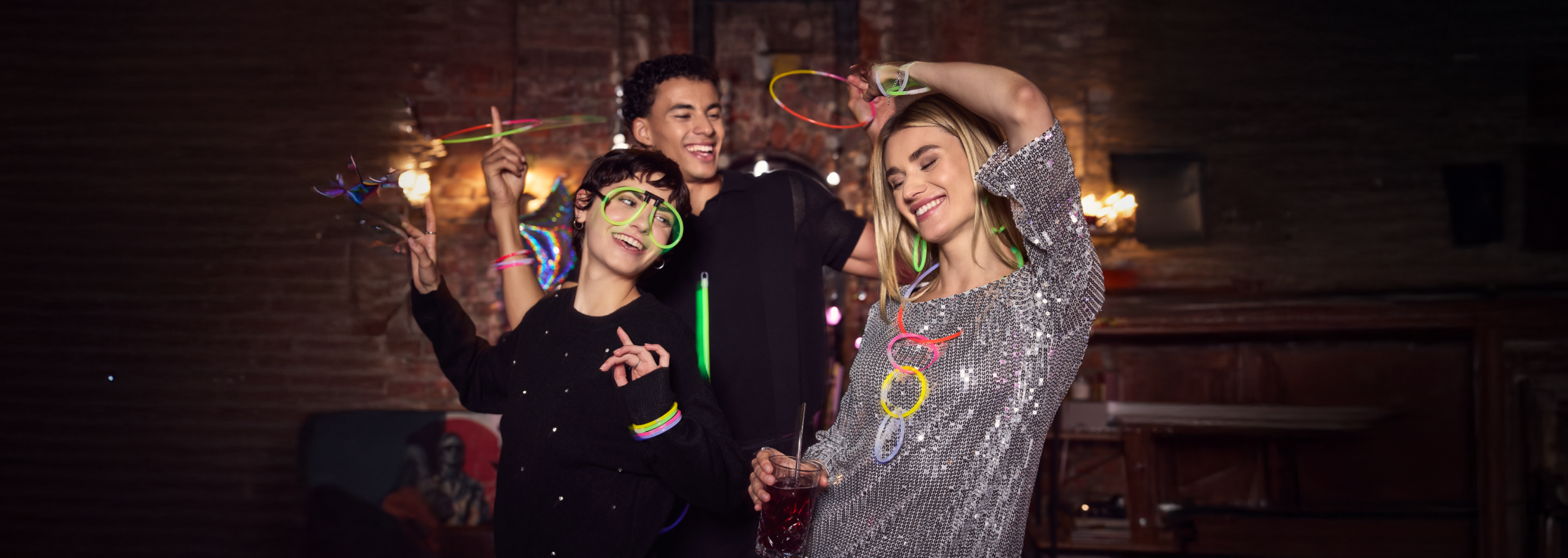 Três amigos a dançar numa festa com óculos e acessórios que brilham no escuro.