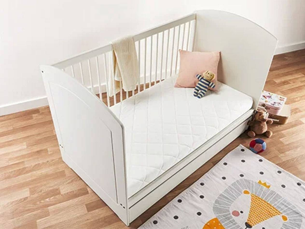 Berço branco para bebé com colchão, almofada e brinquedos num chão de madeira.