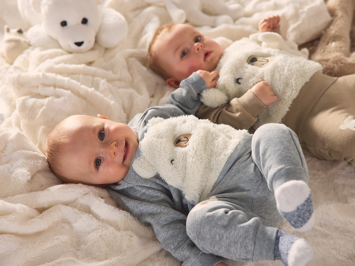 Dois bebés deitados numa manta fofa, vestidos com fatos de urso de peluche.
