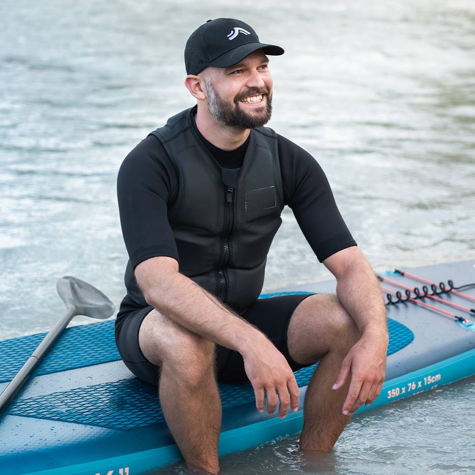 Homem sorridente com colete salva-vidas e boné, sentado numa prancha de paddleboard na água.