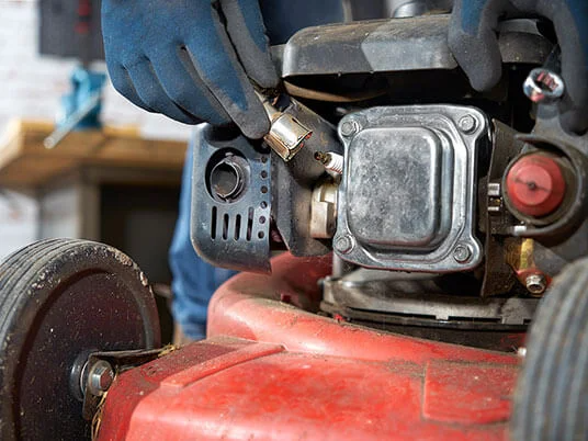 Mãos enluvadas substituindo a vela de ignição de um cortador de grama vermelho.