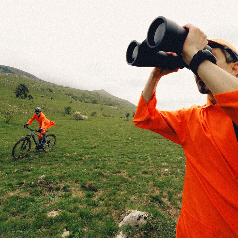 Homem com binóculos e ciclista em roupa desportiva laranja numa paisagem montanhosa.