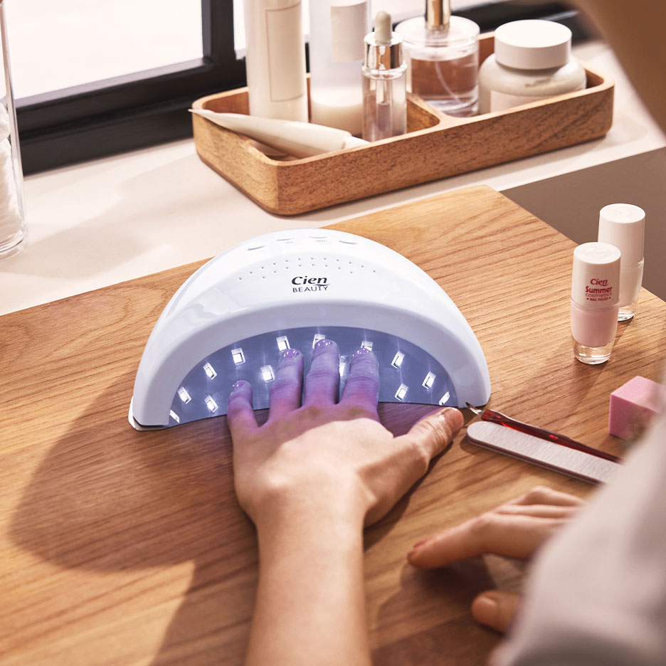 Mulher a secar as unhas numa lâmpada UV, com vernizes e acessórios de manicure por perto.