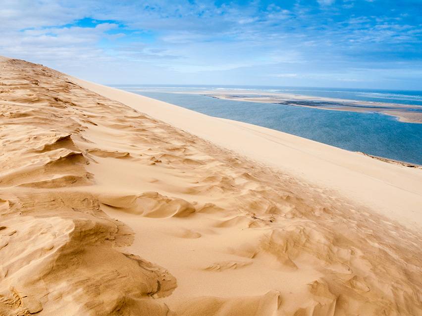 Duna de areia dourada com uma lagoa azul e céu nublado ao fundo.