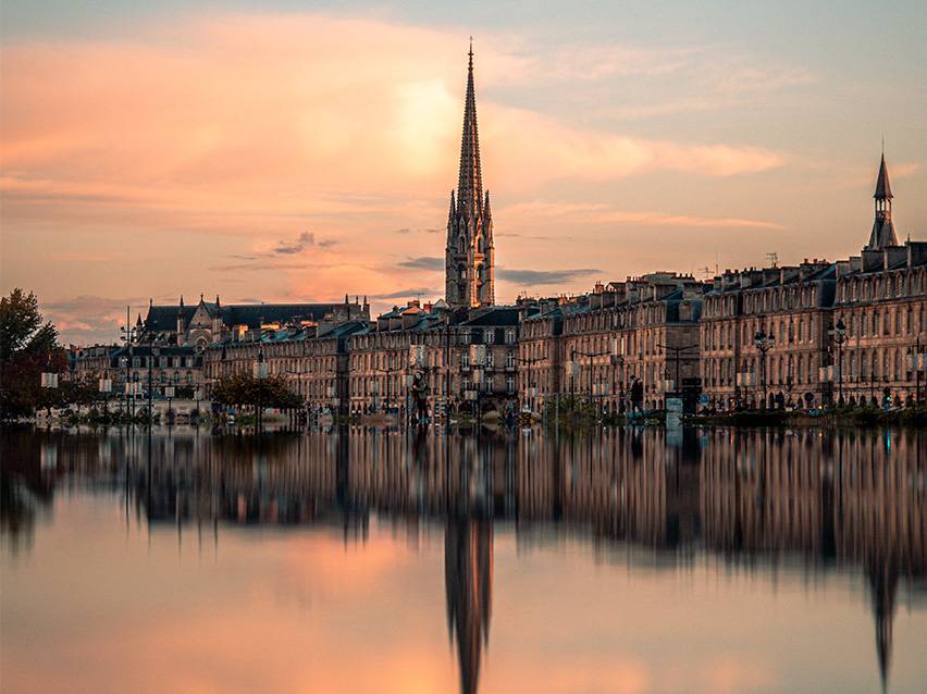 Paisagem urbana de Bordéus ao pôr do sol com uma igreja e o seu reflexo na água.