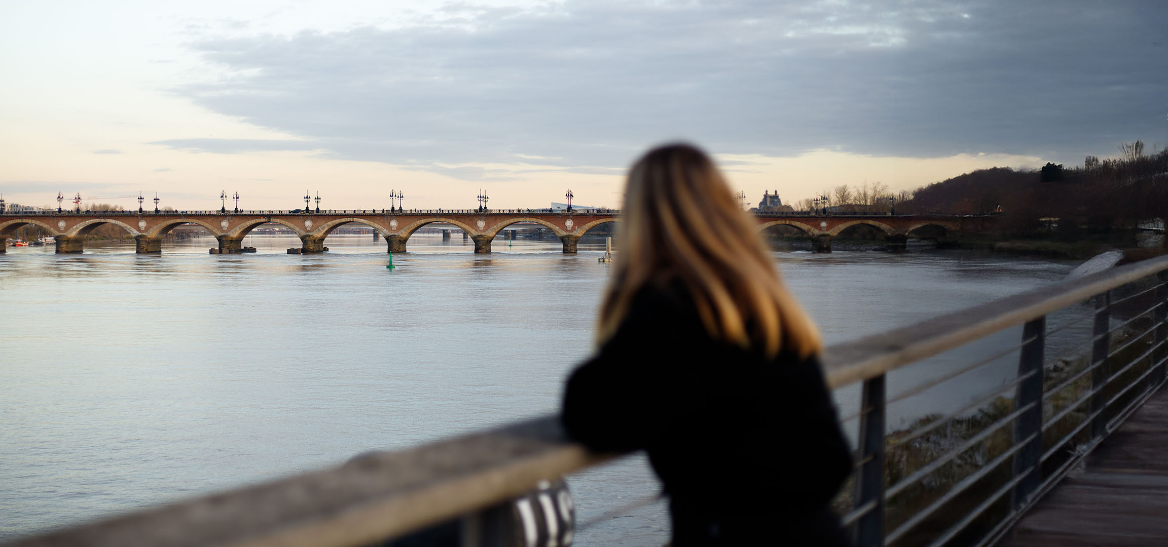 Vista de uma ponte em arco e um rio, com uma pessoa de costas e cabelo comprido.