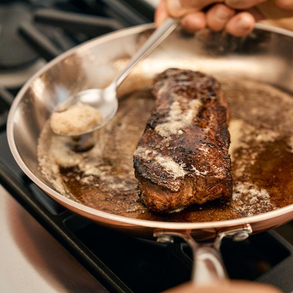 Um bife a cozinhar numa frigideira com manteiga a ser regada por cima.