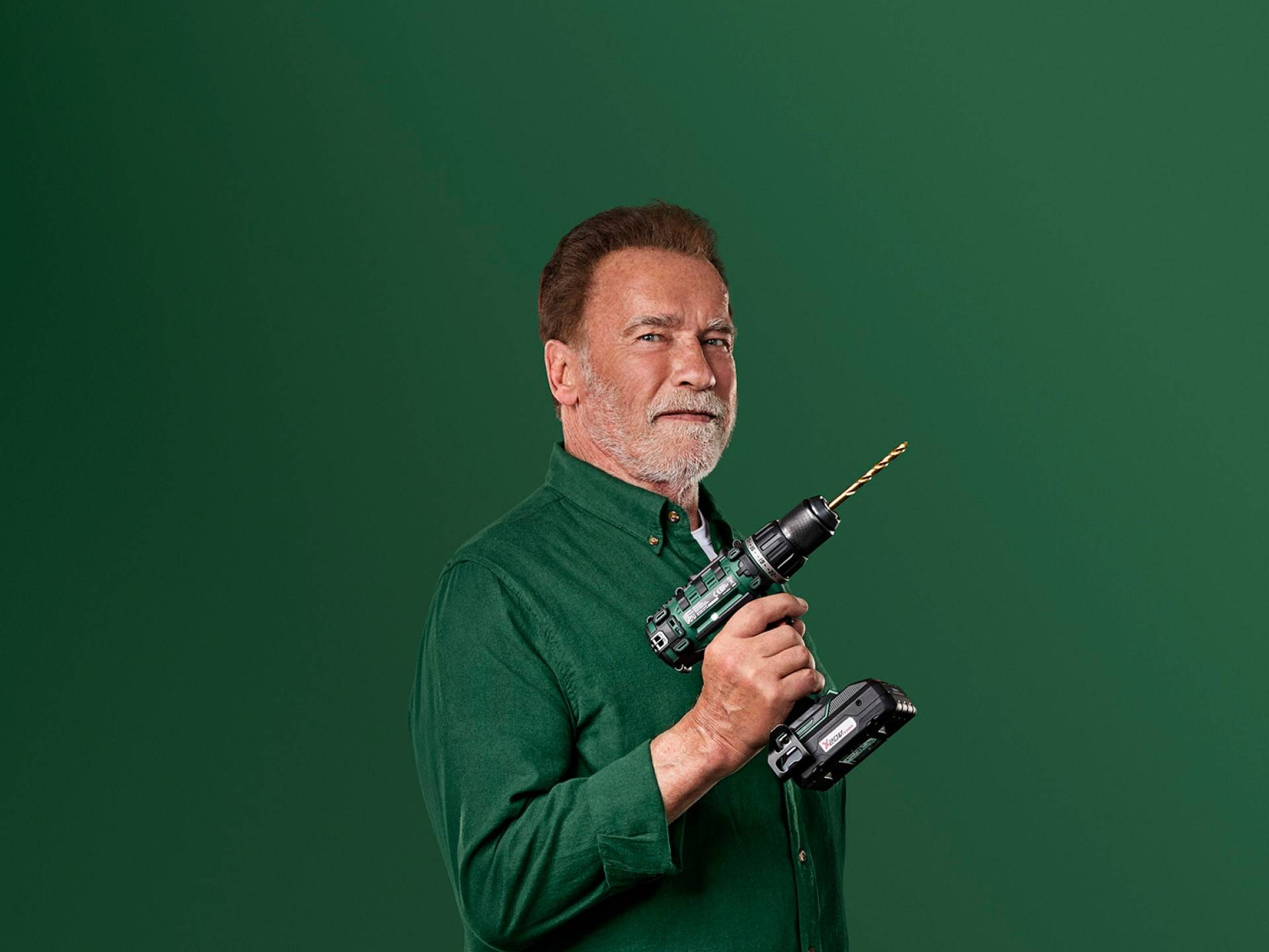 Arnold Schwarzenegger segura uma furadeira elétrica verde em um fundo verde.