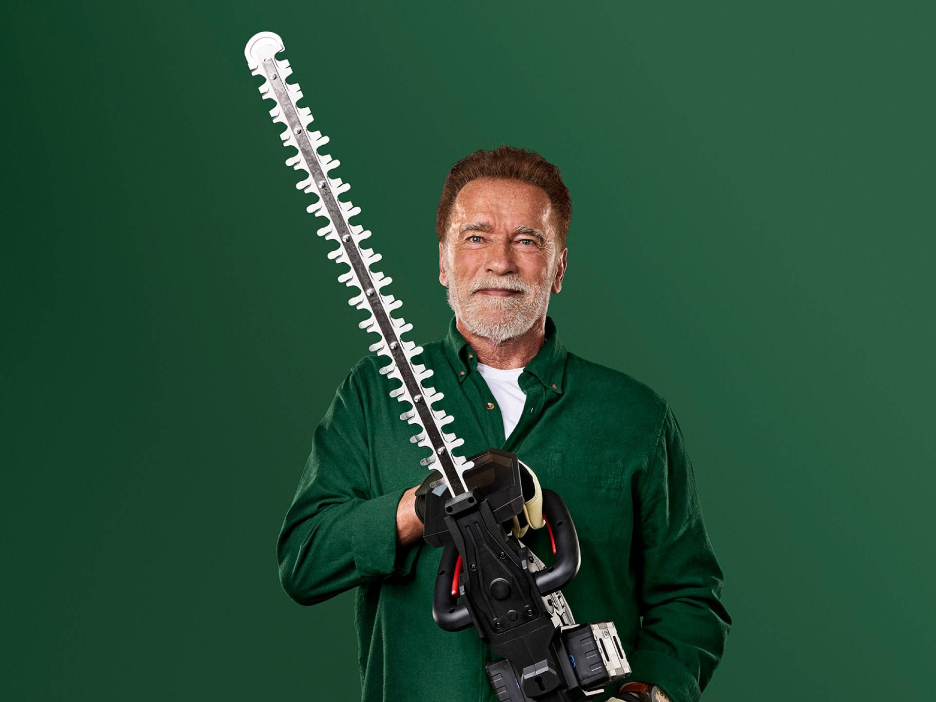 Arnold Schwarzenegger segura um grande aparador de sebes contra um fundo verde.