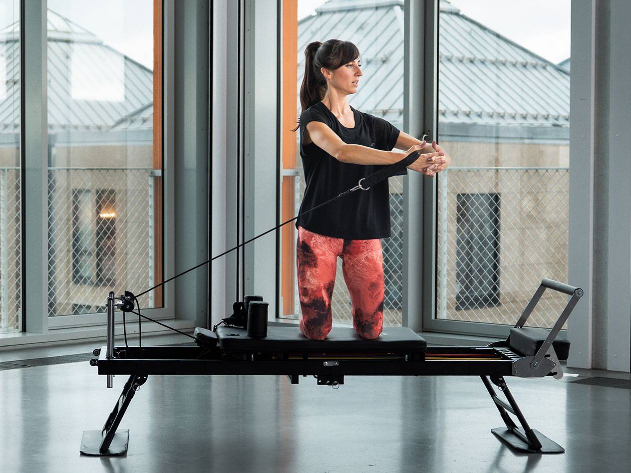 Mulher a fazer Pilates numa máquina reformer preta num quarto luminoso.