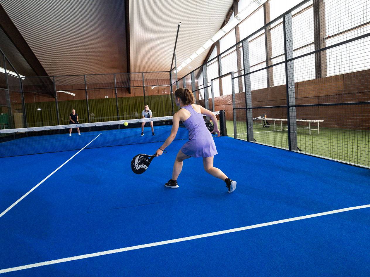 Mulher a jogar padel num campo azul, com outros jogadores ao fundo.