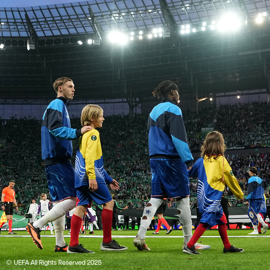 Jogadores de futebol e crianças caminhando no campo de um estádio lotado.