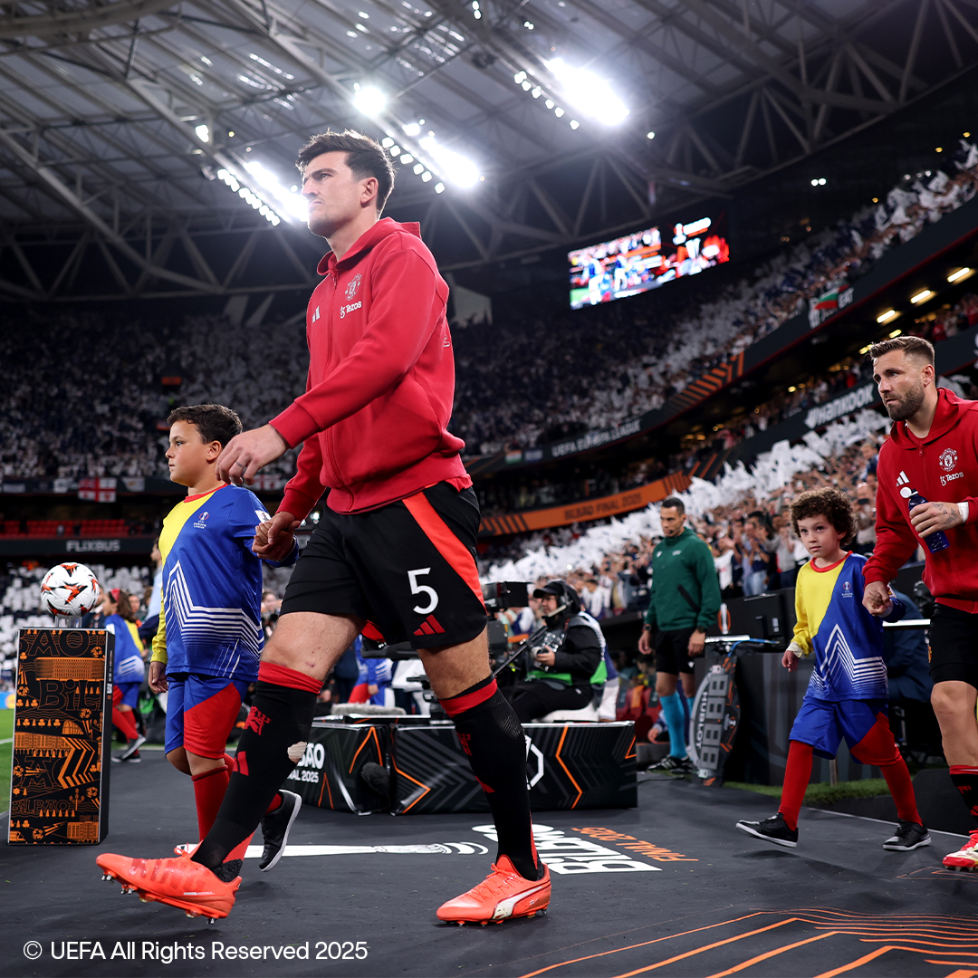 Jogadores de futebol com camisas vermelhas e calções pretos caminham em campo com crianças, vestindo roupas esportivas.