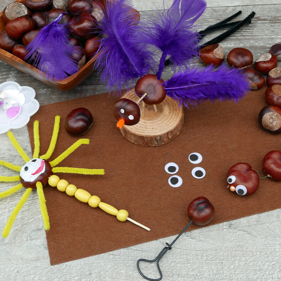 Artesanato de castanhas com penas roxas, olhos móveis e arames felpudos numa mesa de madeira.