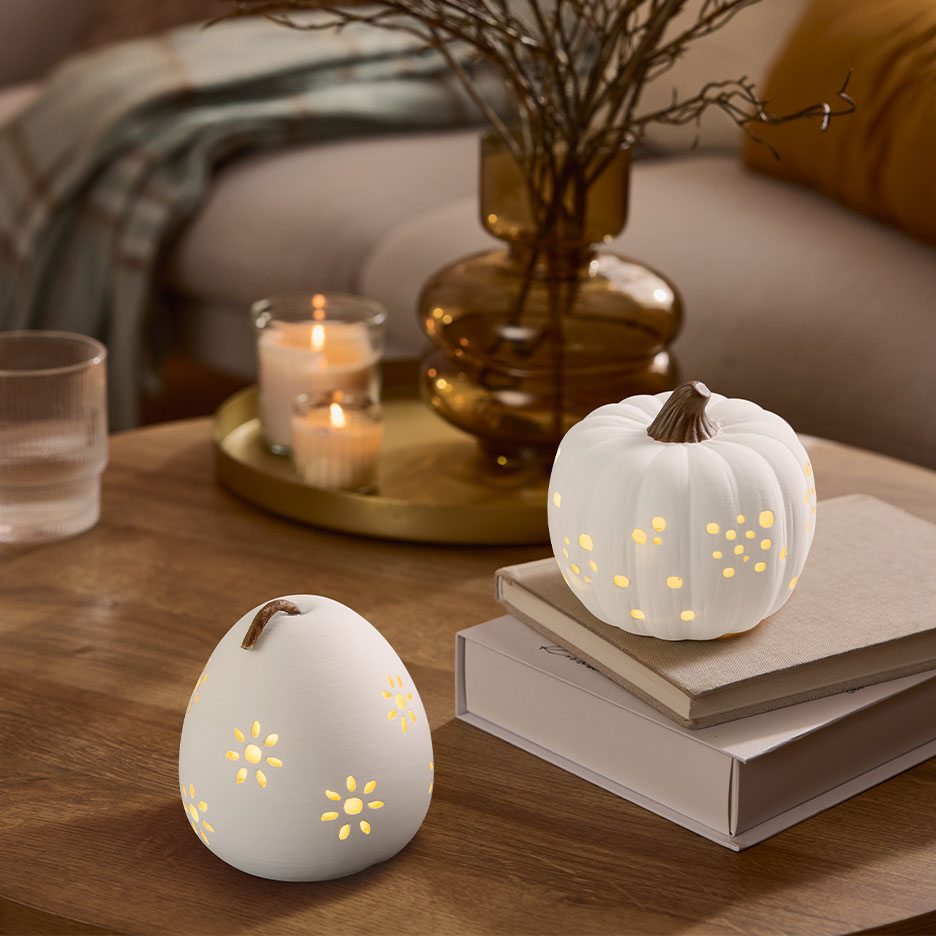 Duas abóboras decorativas brancas iluminadas em uma mesa de madeira com velas e um vaso.