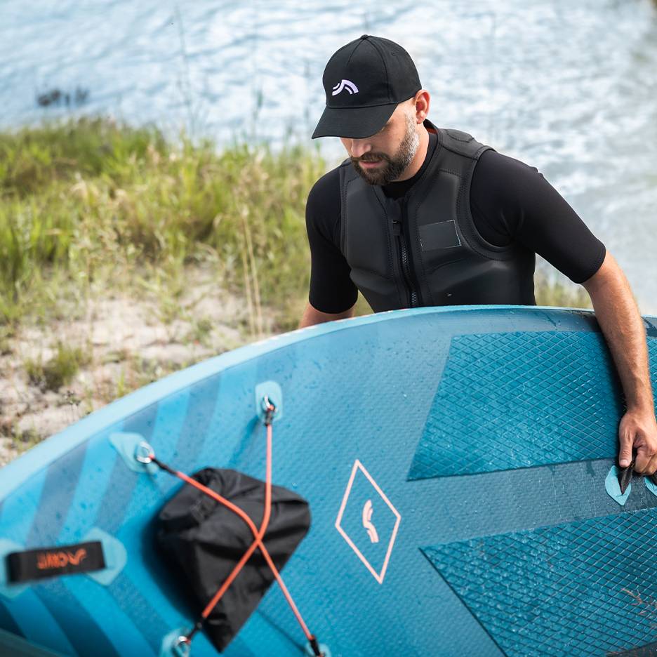 Homem com colete salva-vidas e boné, a segurar uma prancha de paddleboard azul junto à água.