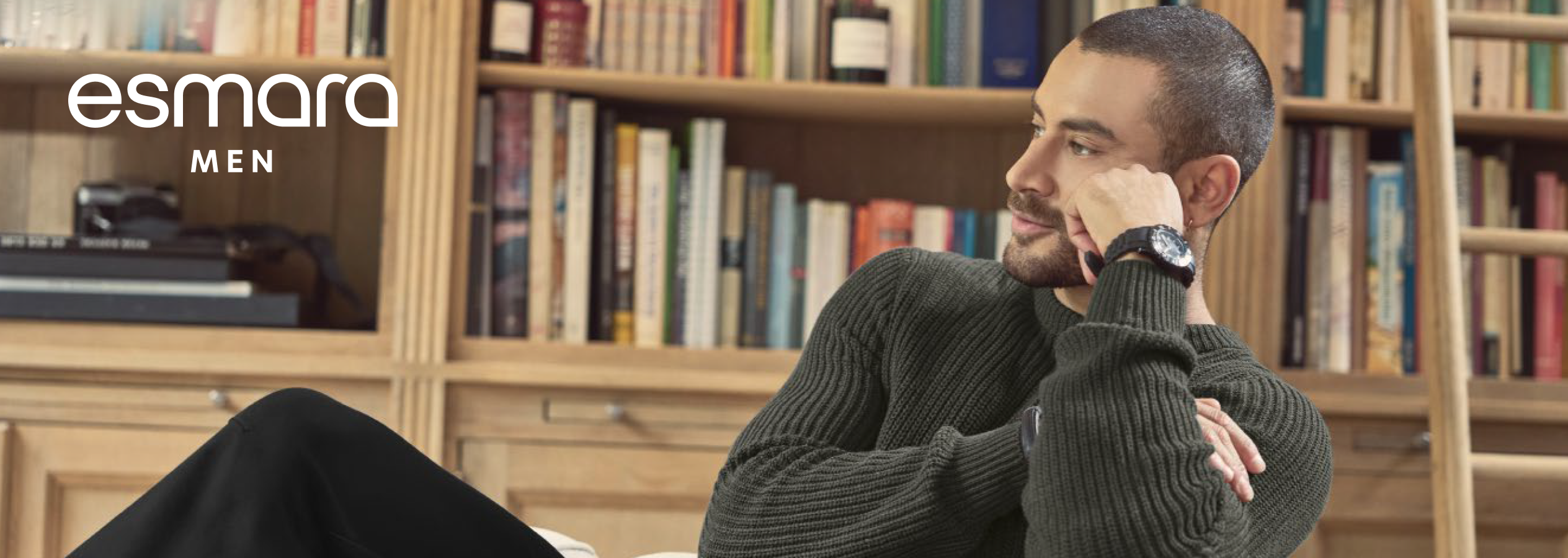 Homem com camisola de malha Esmara Men, sentado em frente a uma estante de livros.