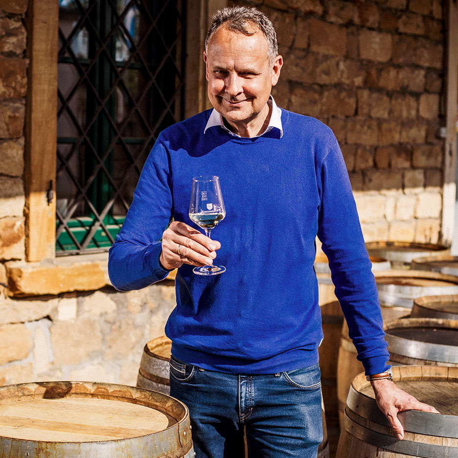 Homem sorridente de camisola azul a segurar um copo de vinho, com barris de madeira ao fundo.
