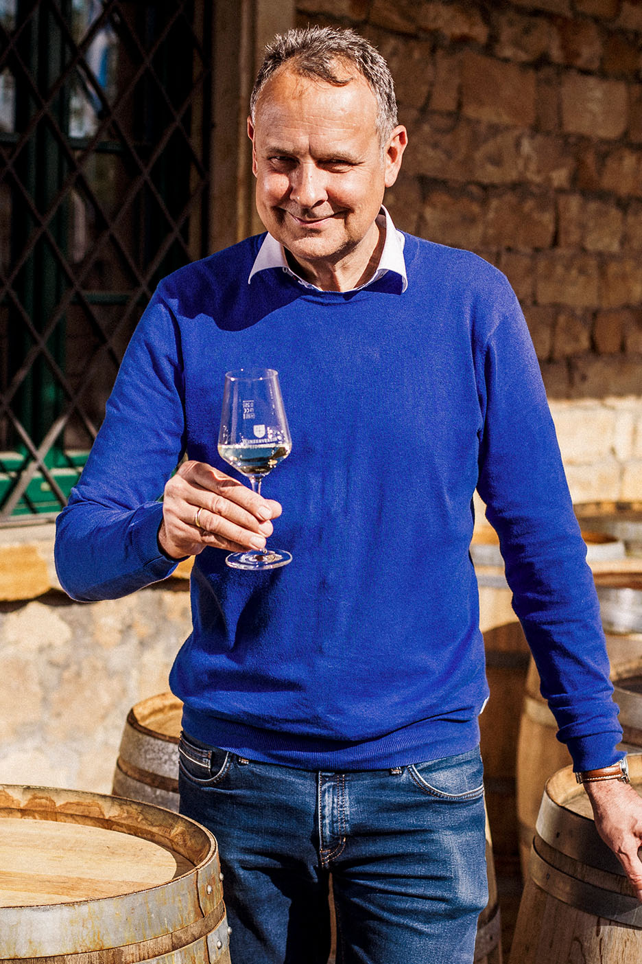 Homem sorridente de camisola azul e jeans, segurando um copo de vinho branco entre barris de madeira.
