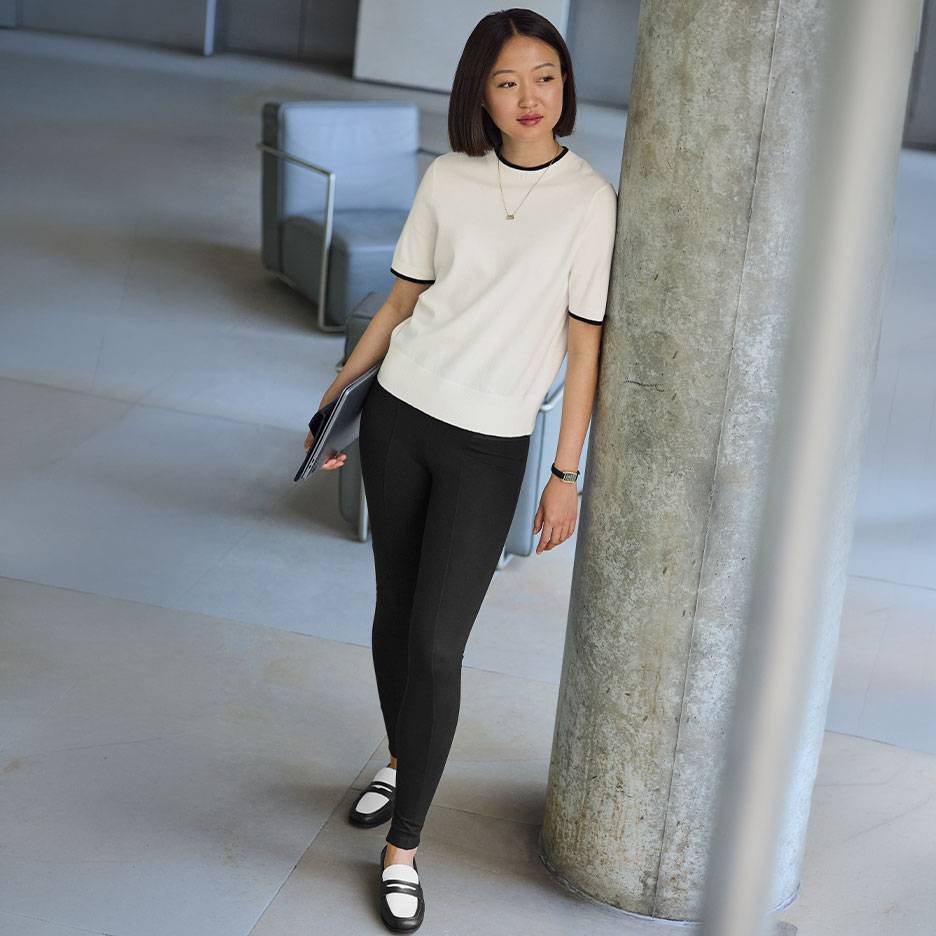 Mulher com top branco de manga curta com acabamento preto, leggings pretas e mocassins bicolores.