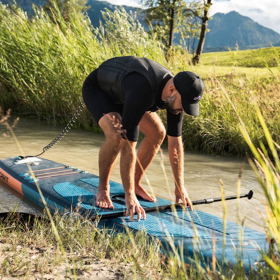 Homem em fato de mergulho e colete de flutuação a preparar-se para stand-up paddle num rio.