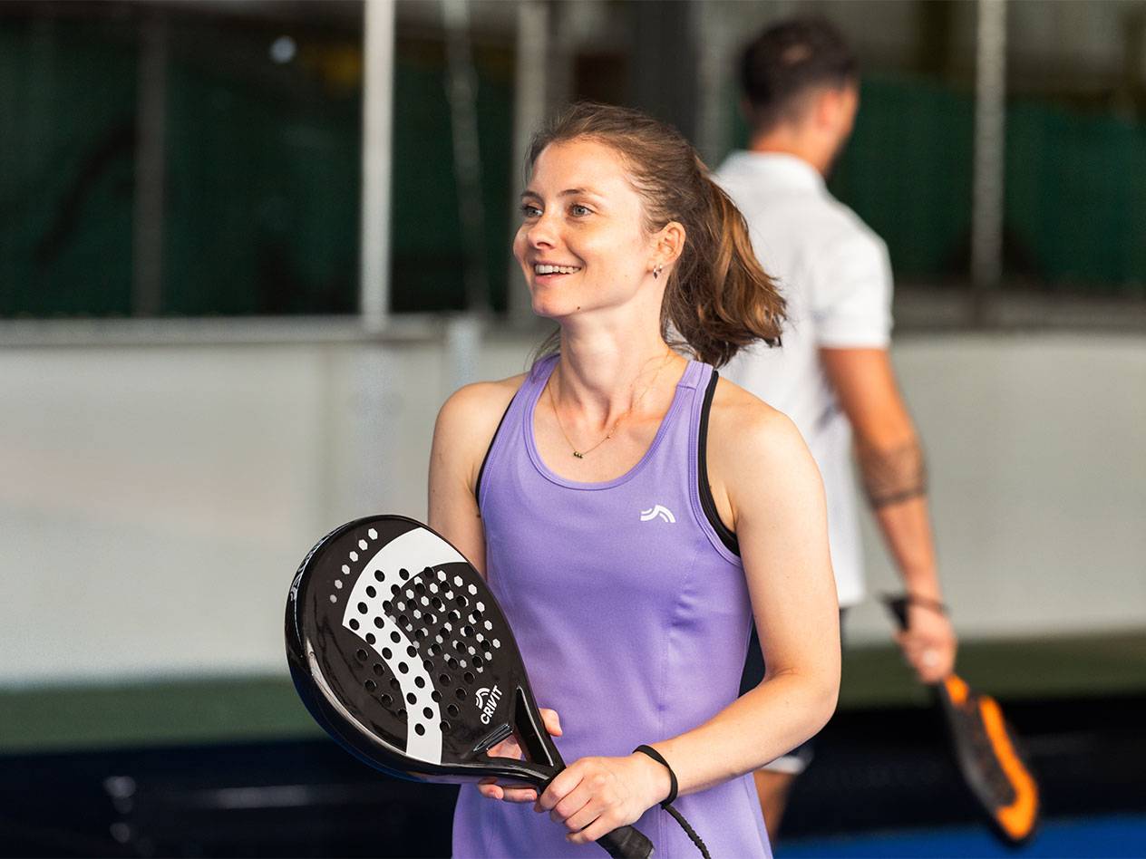 Mulher de vestido desportivo roxo segurando uma raquete de padel, homem ao fundo.