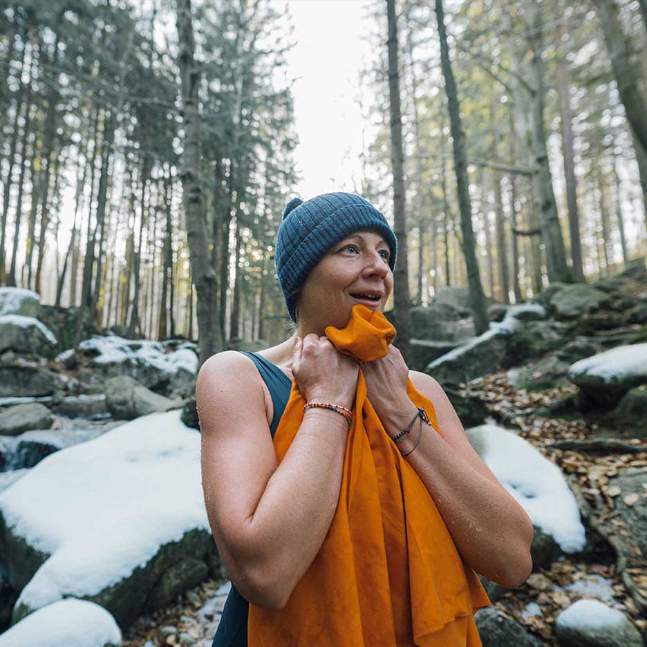 Mulher de gorro e fato de banho a aquecer-se com uma toalha numa floresta nevada.