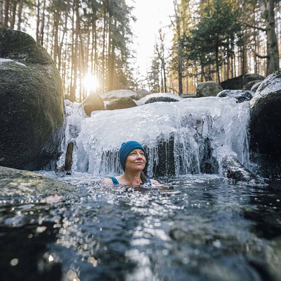 Mulher de gorro e fato de banho azul a desfrutar de um mergulho frio num rio gelado.