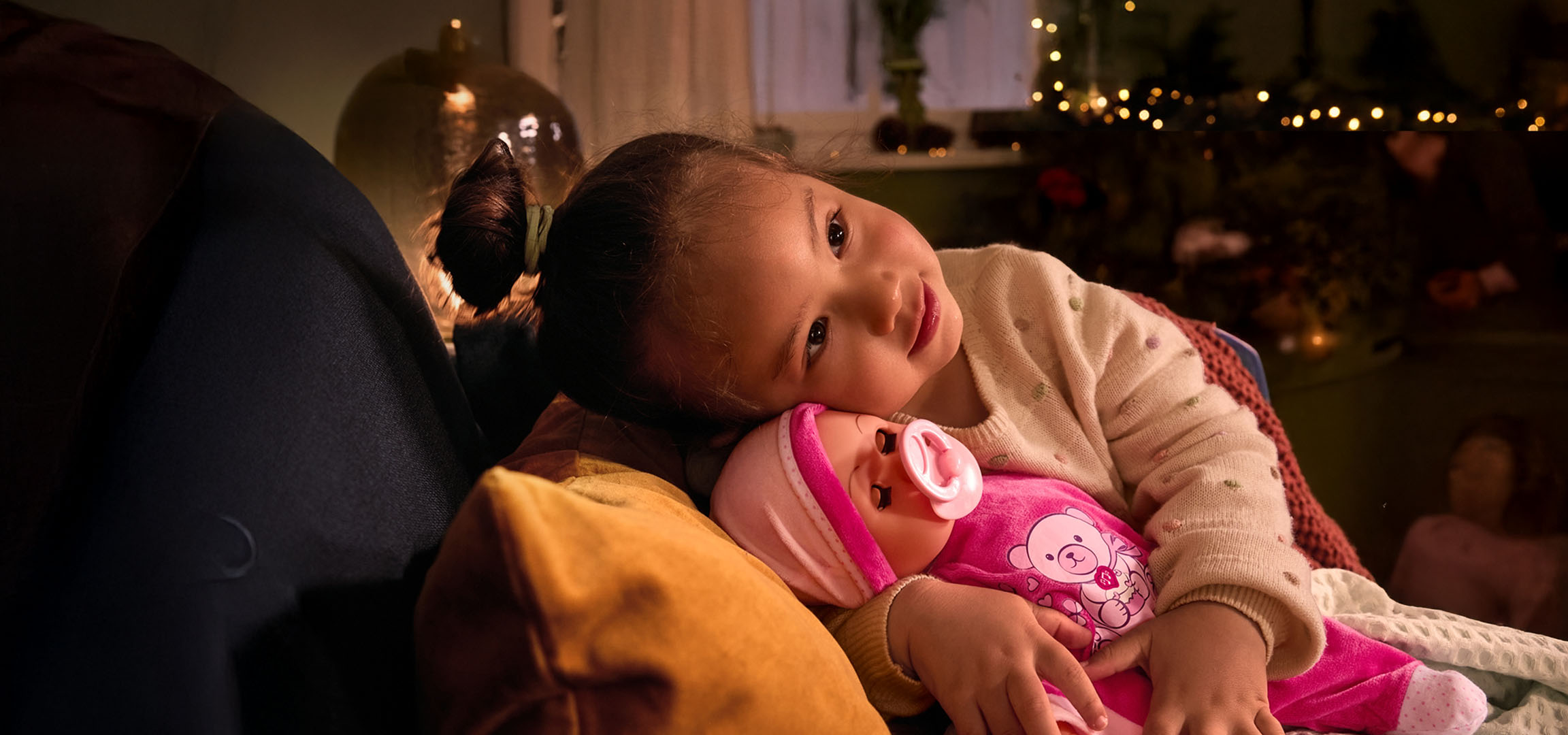 Menina com boneca e chupeta, aconchegada numa almofada.