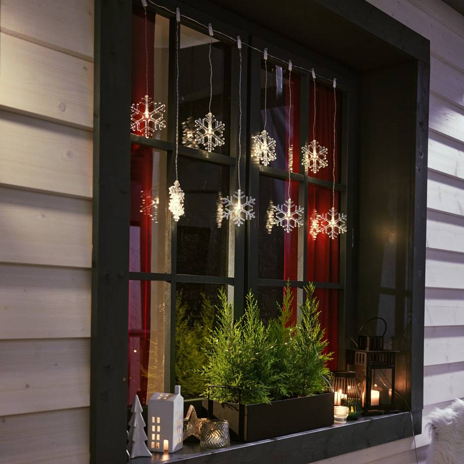 Luzes de floco de neve penduradas numa janela com pequenos pinheiros e velas no parapeito.