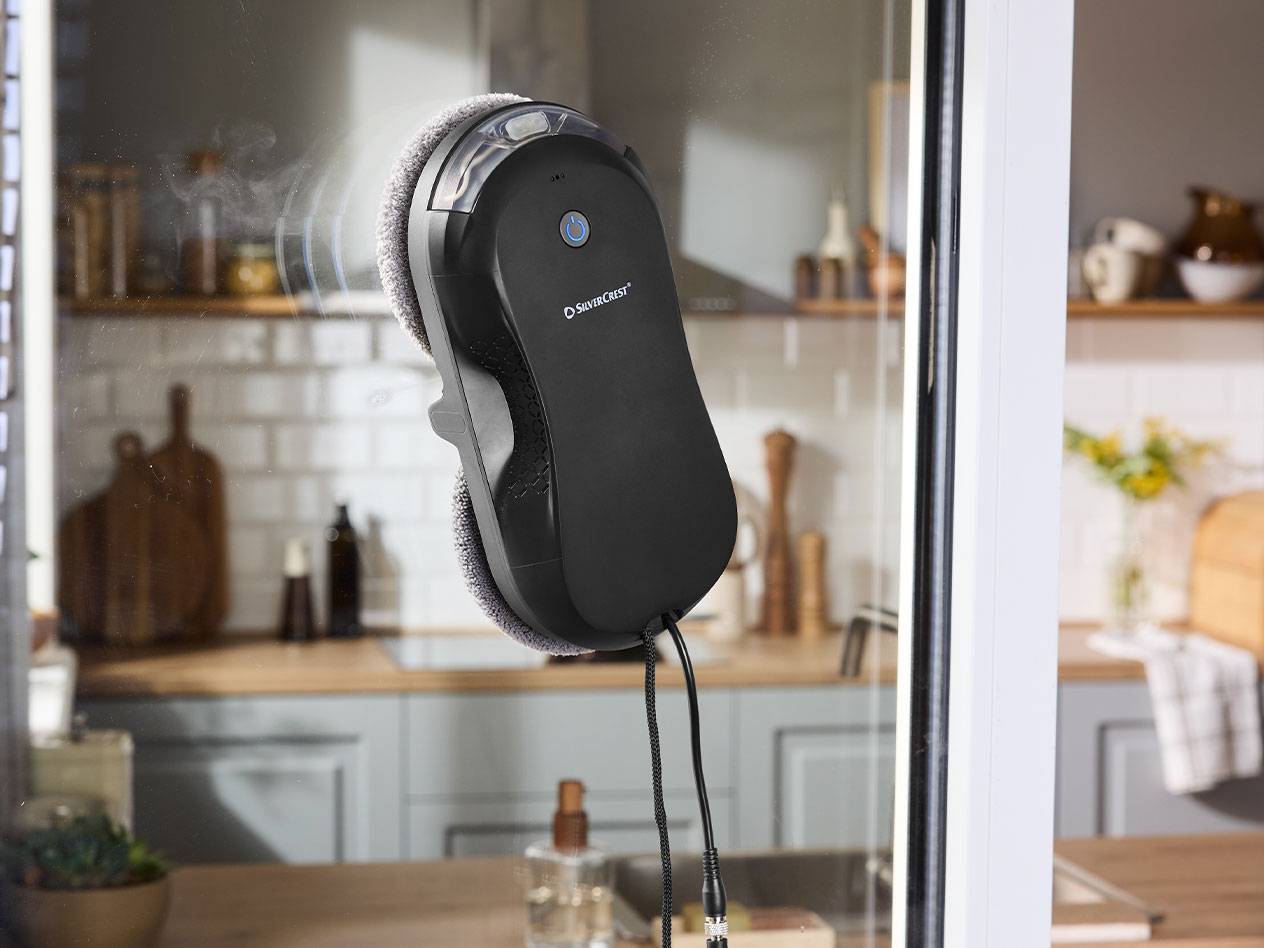 Robô de limpeza de janelas Silvercrest numa vidraça com uma cozinha ao fundo.