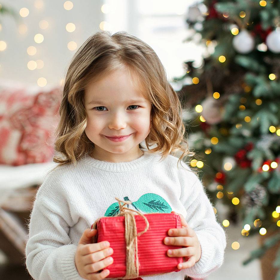 Menina sorridente com um presente de Natal, com uma árvore de Natal iluminada ao fundo.