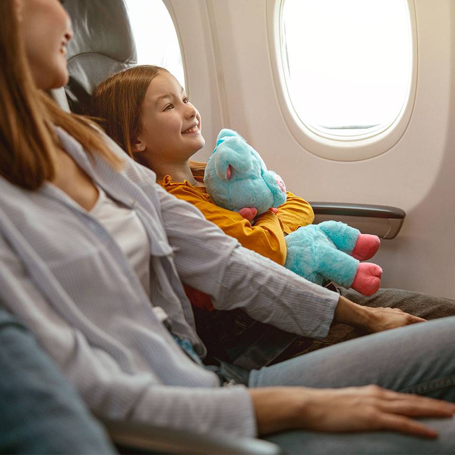 Uma criança com um brinquedo de pelúcia a olhar pela janela de um avião ao lado de um adulto.