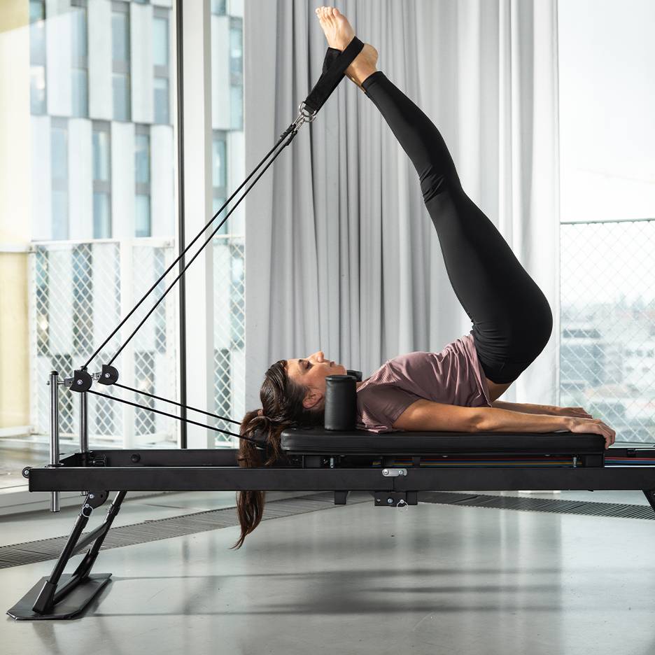 Mulher a fazer Pilates numa máquina preta, com as pernas para cima.