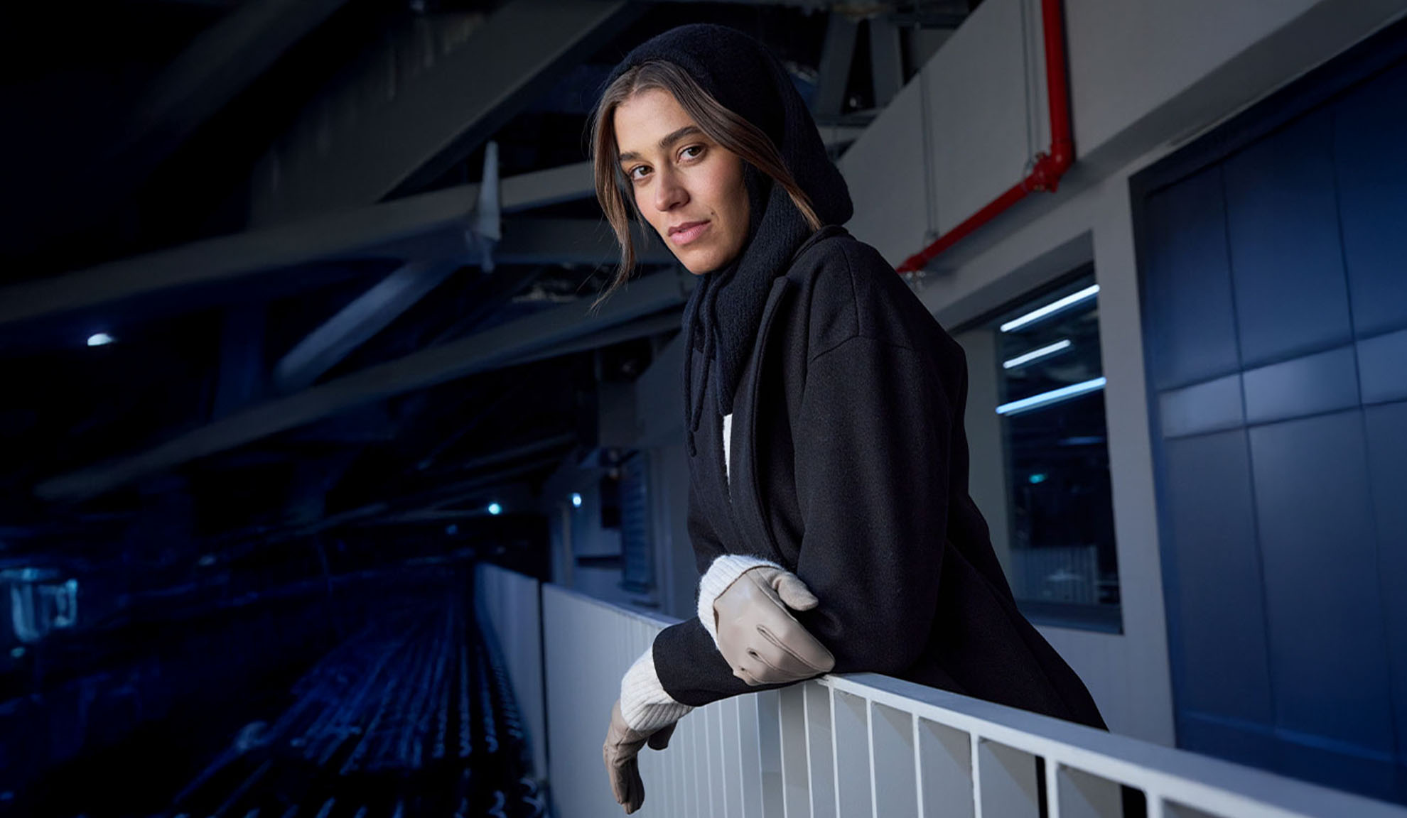 Mulher de casaco preto e cachecol com capuz, apoiada numa grade num ambiente moderno.