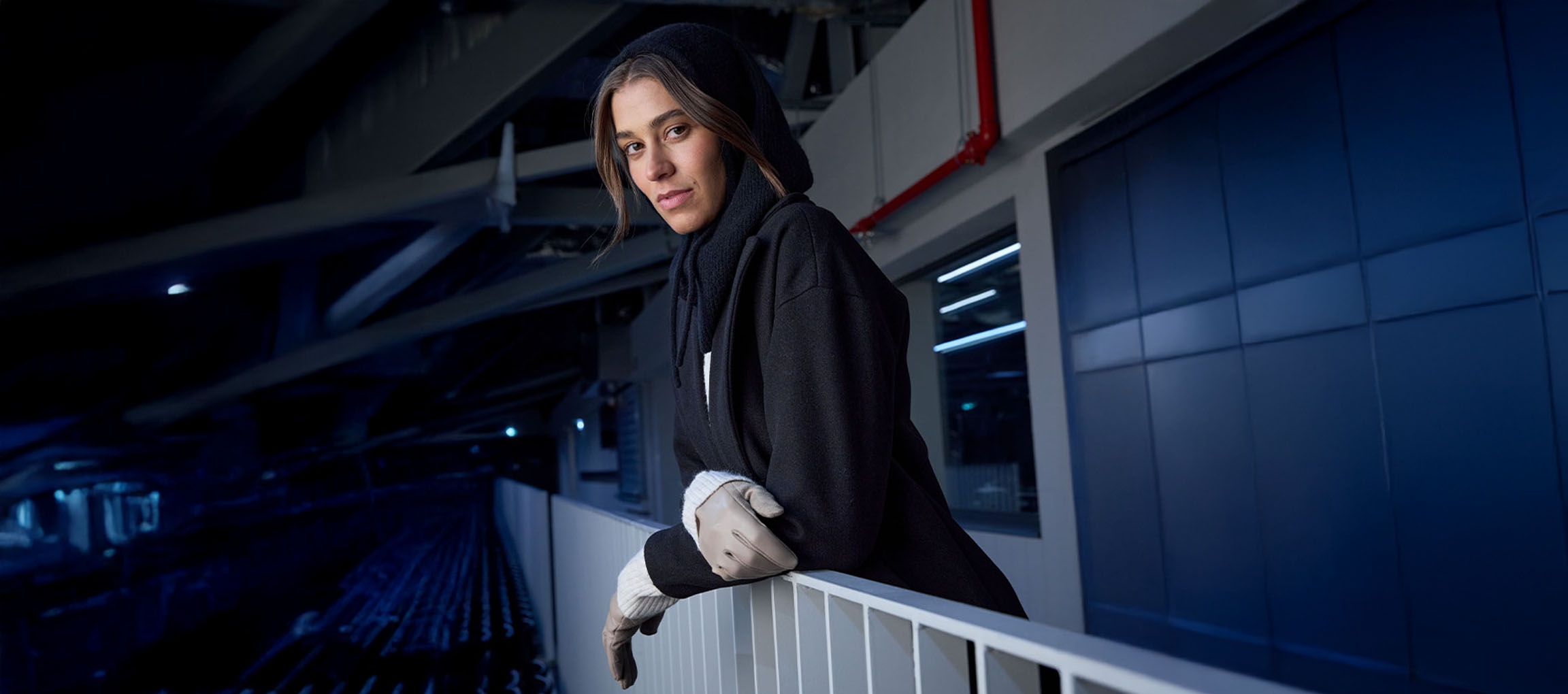 Mulher de casaco preto e luvas encostada a um corrimão em ambiente urbano.