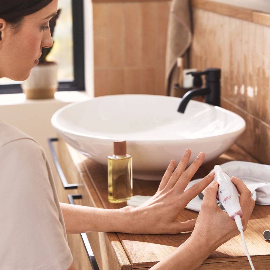 Mulher a usar um conjunto elétrico de manicure/pedicure numa bancada de madeira com lavatório branco.