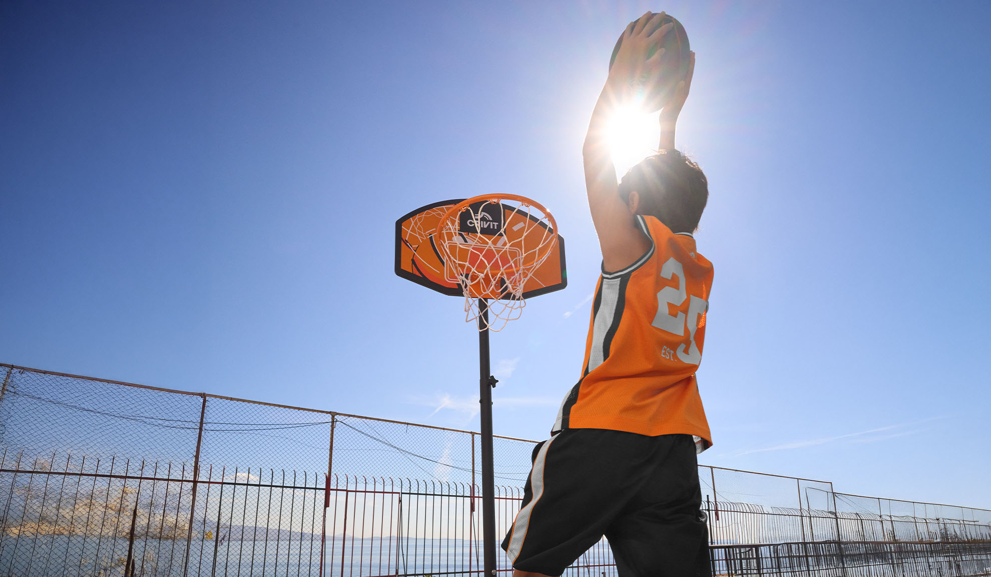 Rapaz com camisola de basquetebol a lançar ao cesto ao ar livre sob um sol forte.