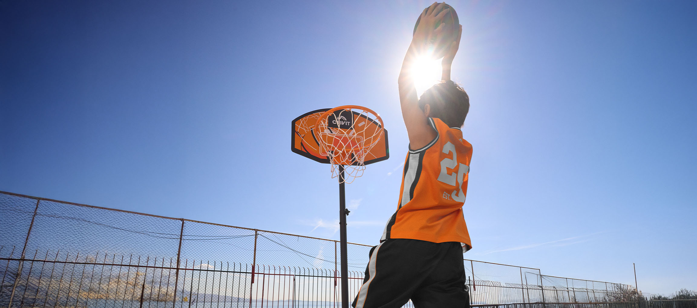 Rapaz com camisola de basquetebol laranja a lançar a bola ao cesto num dia de sol.