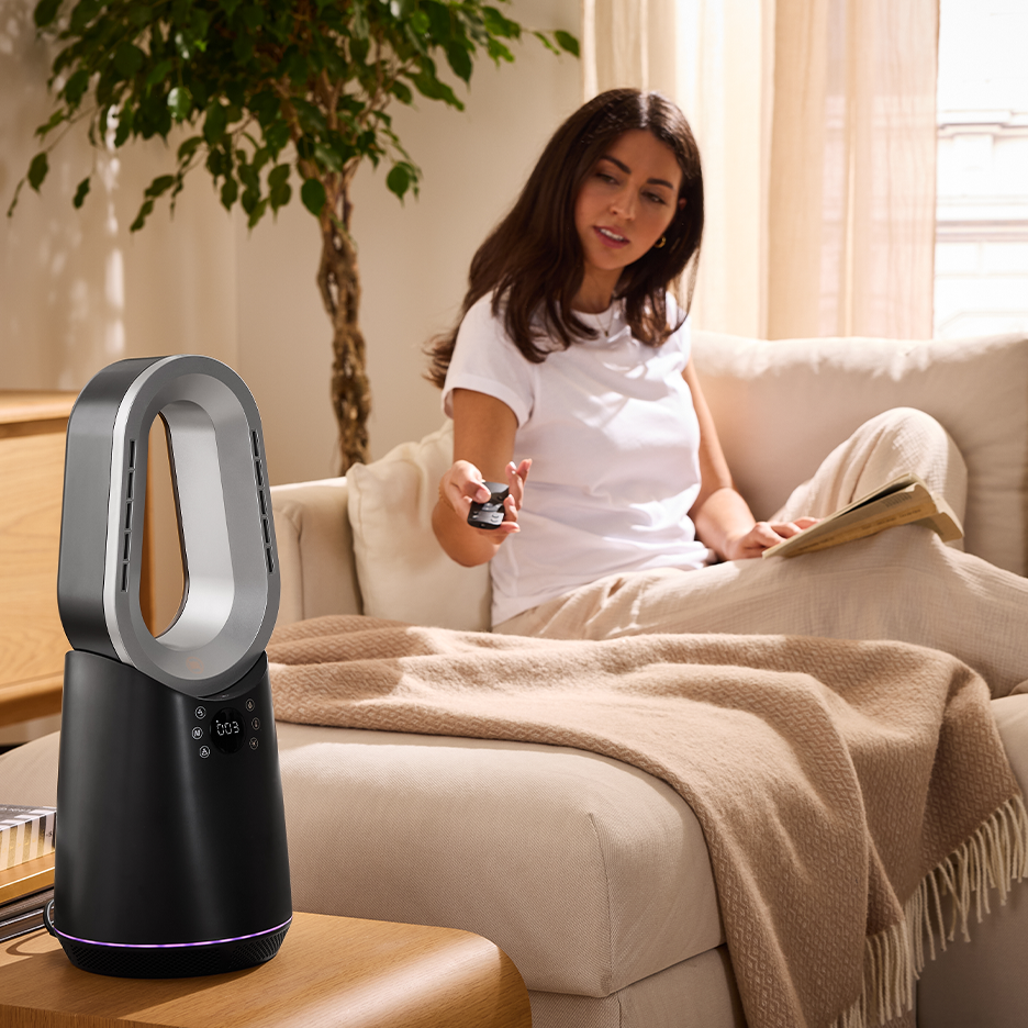 Uma mulher em um sofá usa um controle remoto para controlar um ventilador sem pás preto e prateado.